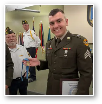 Noah Squires in uniform proudly holds a medal while standing next to another veteran, with additional veterans in the background.